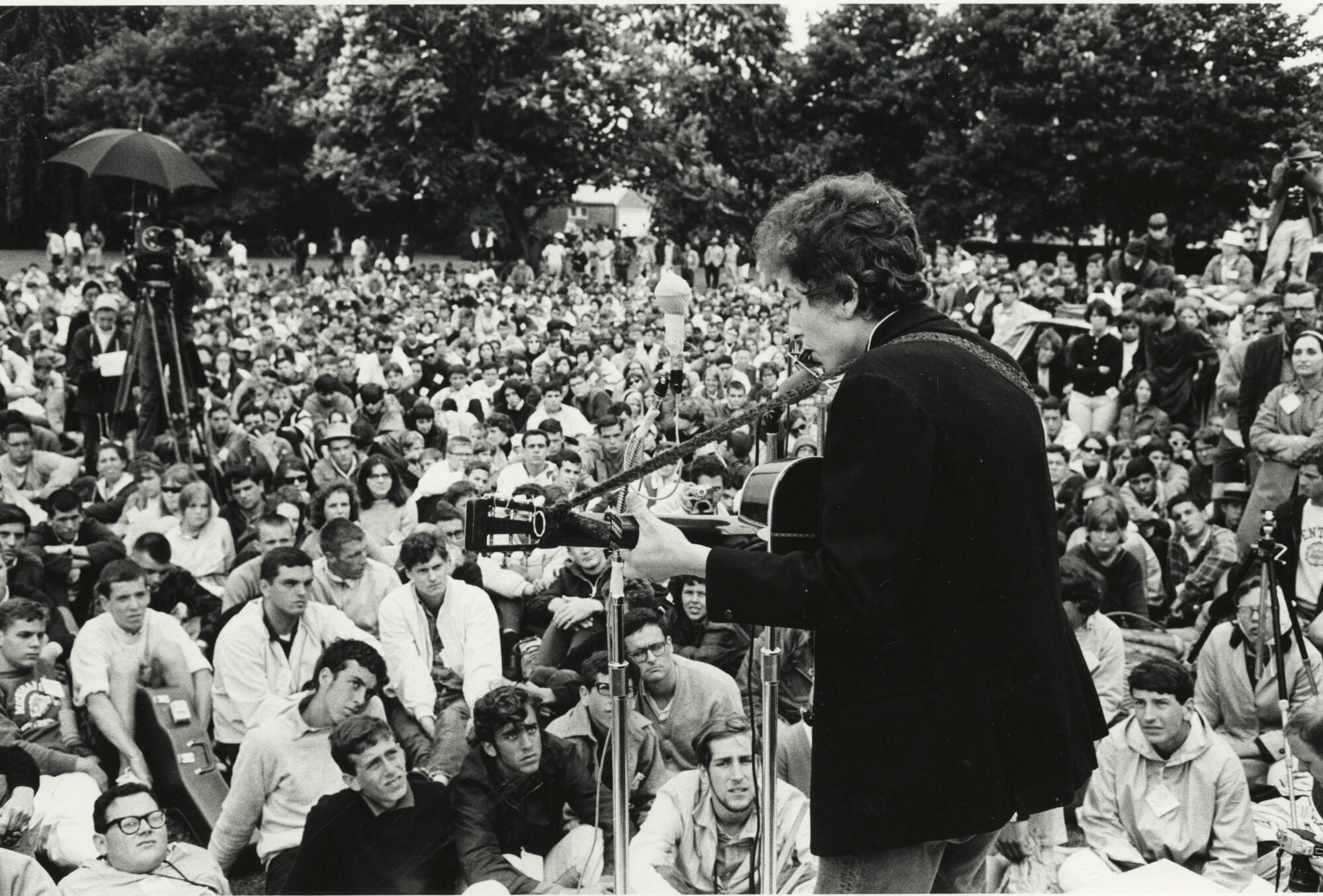 Black and white photograph of Bob Dylan playing a guitar and singing in front of a large crowd outdoors.